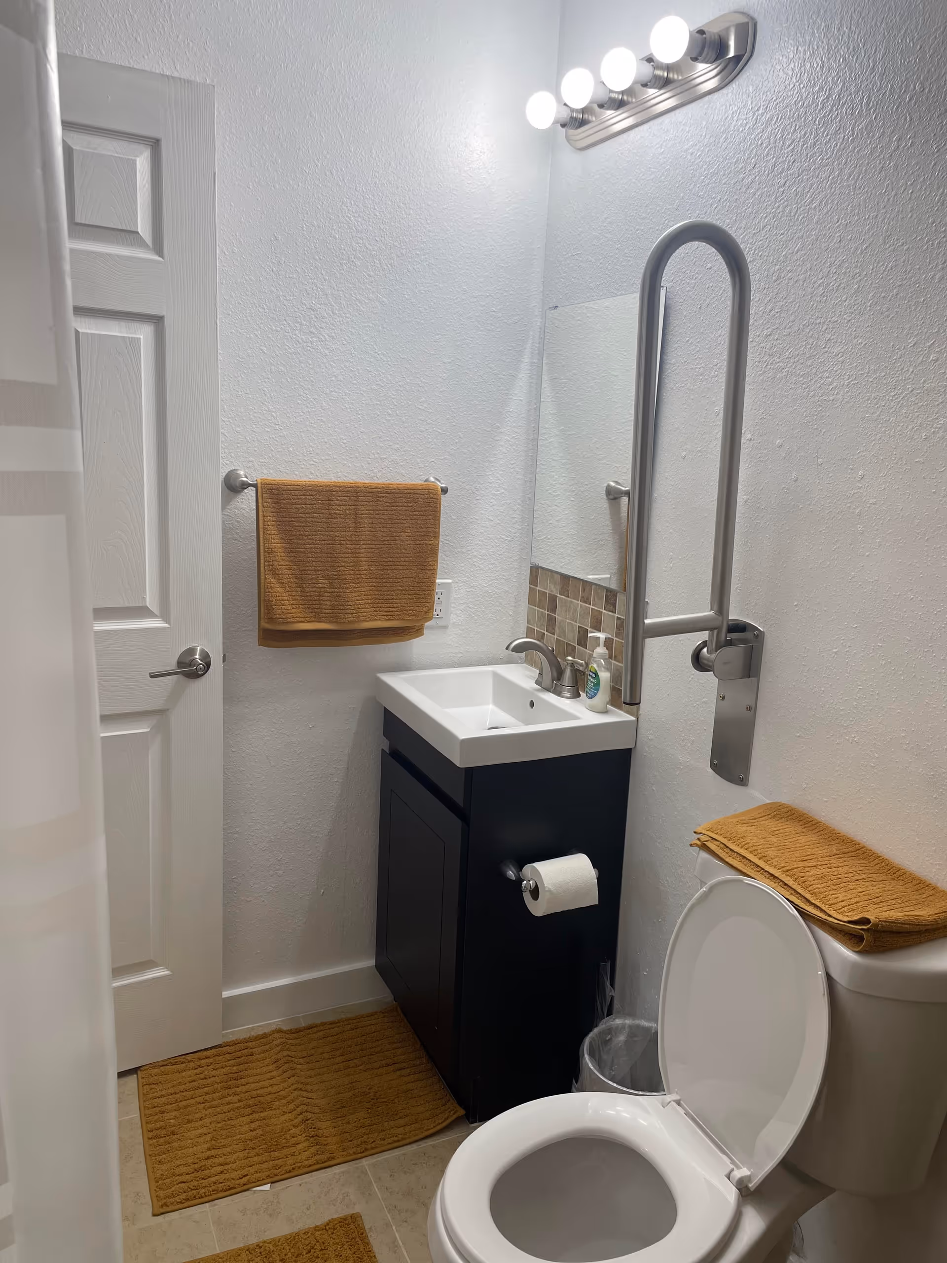 Compact bathroom featuring a dark vanity, toilet, towel bars, grab bar, and beige mats.