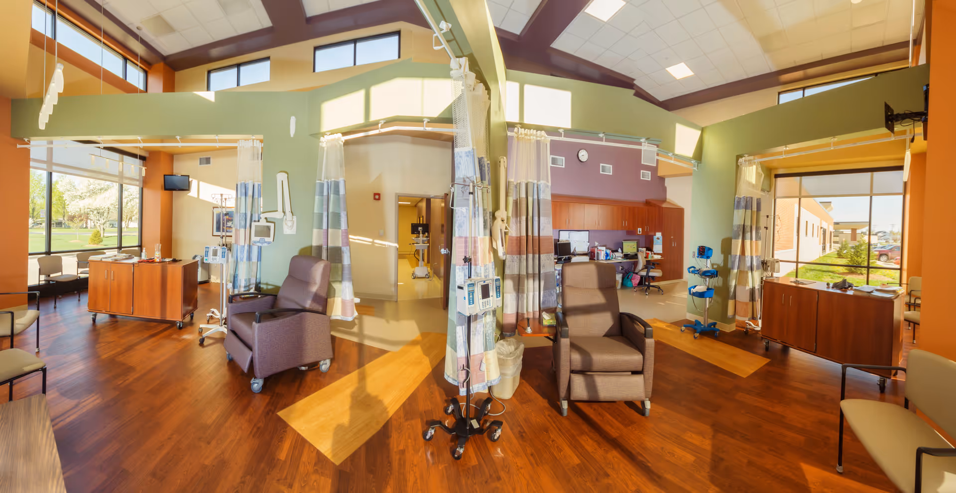 Bright hospital lounge with recliners, privacy curtains, medical equipment, and wood floors.