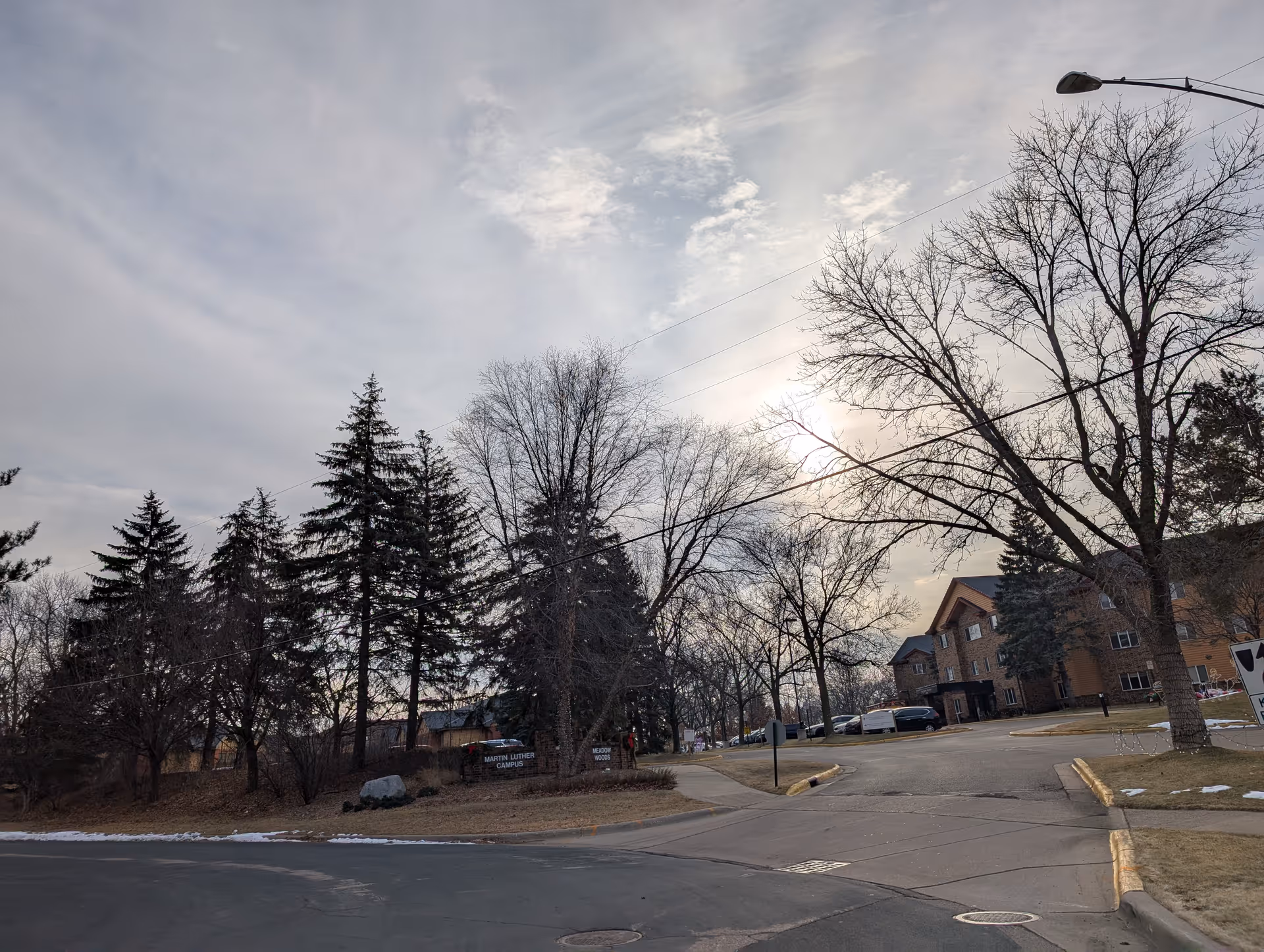 Exterior view of the Meadow Woods Assisted Living entrance with trees, a driveway, and a multi-story brick building under a cloudy sky.