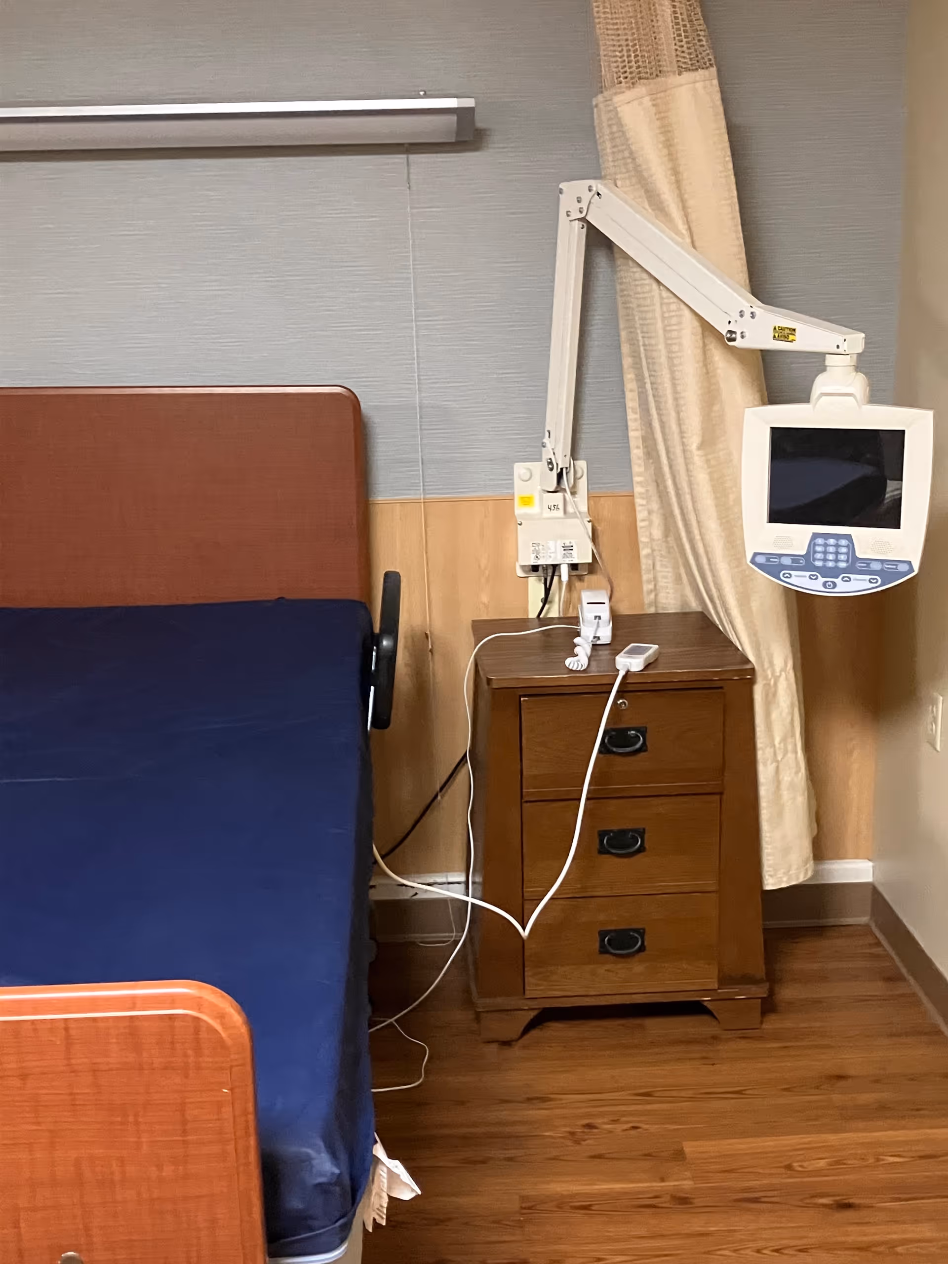 Interior of a patient bedroom with bed, nightstand, and mounted medical monitor.
