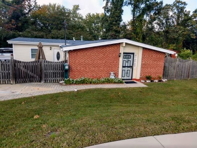 Single-story brick-front house with a fenced yard, front door, mailbox, and grassy lawn in front of trees.