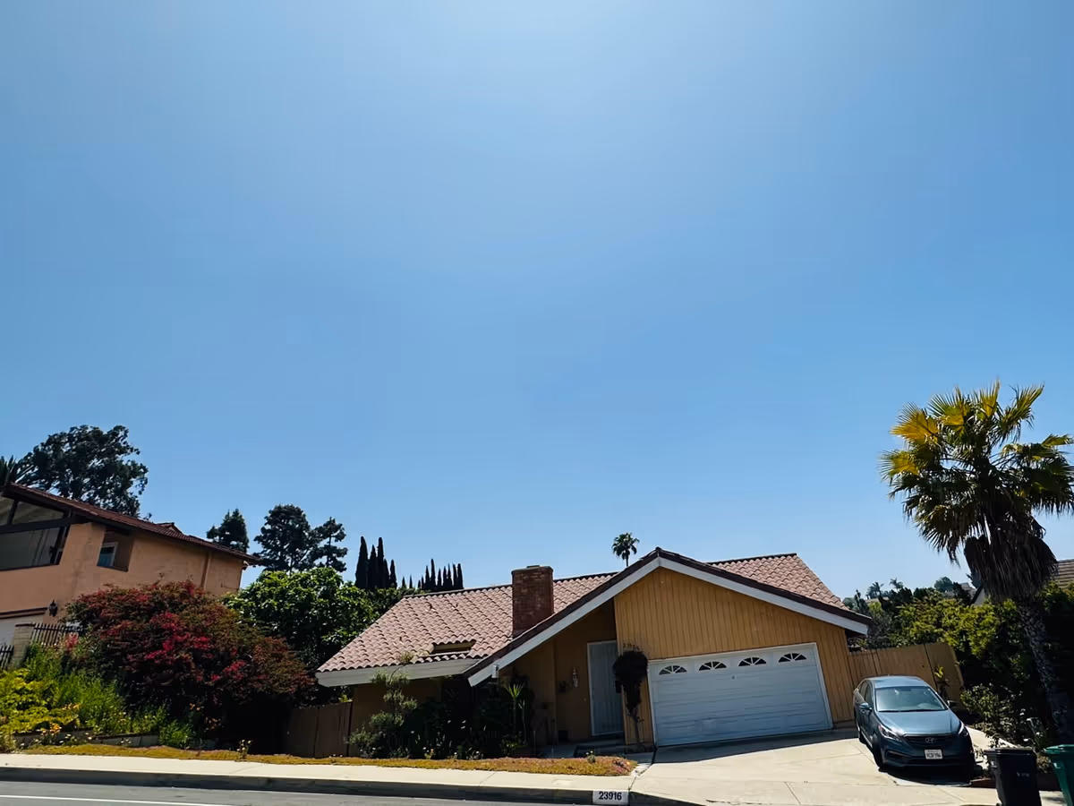Single-story suburban house with a tiled roof, garage, driveway, and palm tree under clear sky.