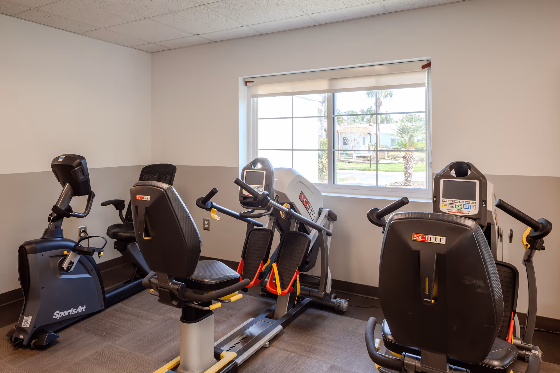 Cardio fitness room with multiple exercise machines by a large window inside a senior care facility.