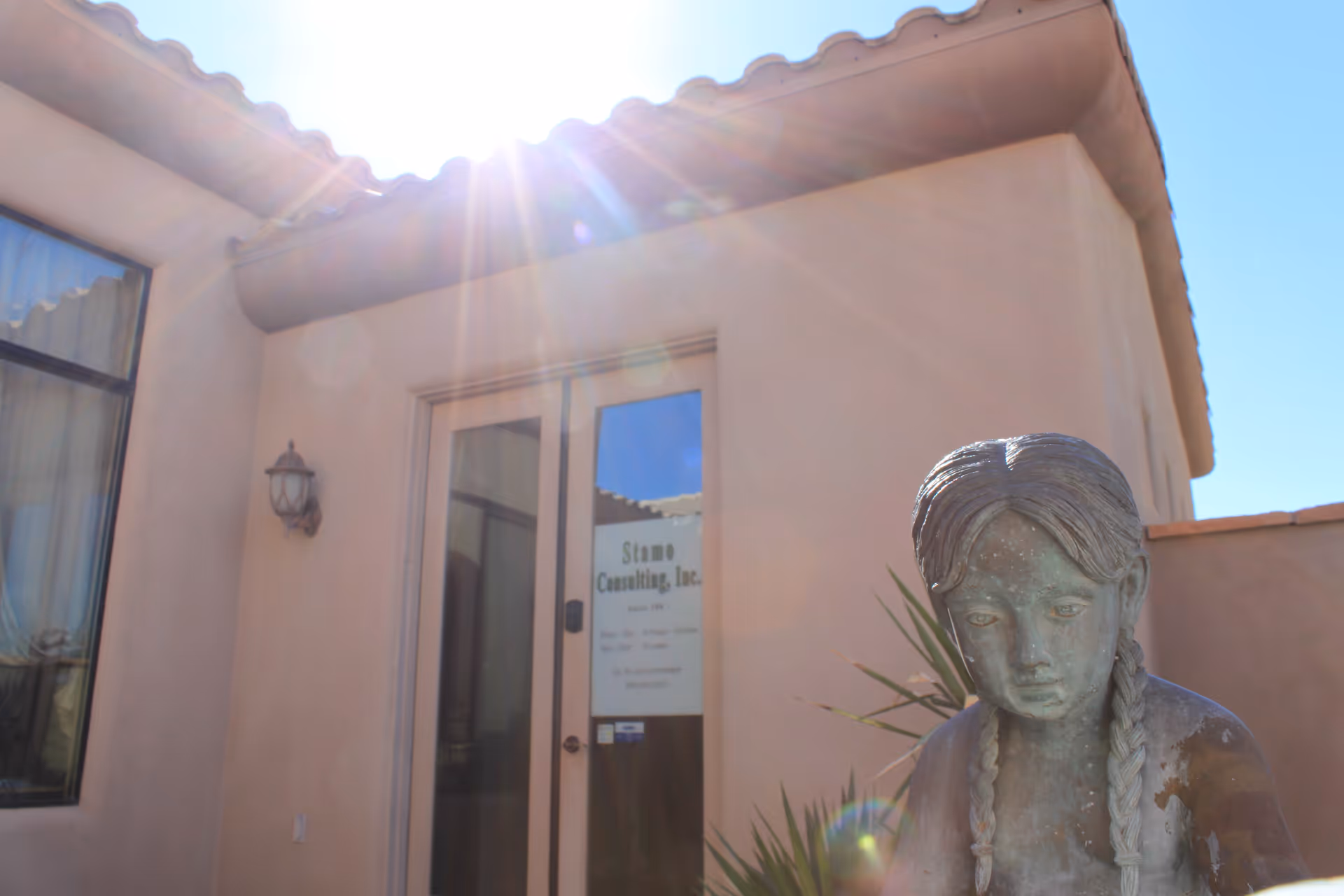 Sunlit stucco building exterior with glass door labeled Stamo Consulting and a nearby statue.