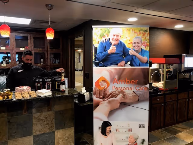 Staff member serves at a service counter beside an Amber Personal Care banner indoors.