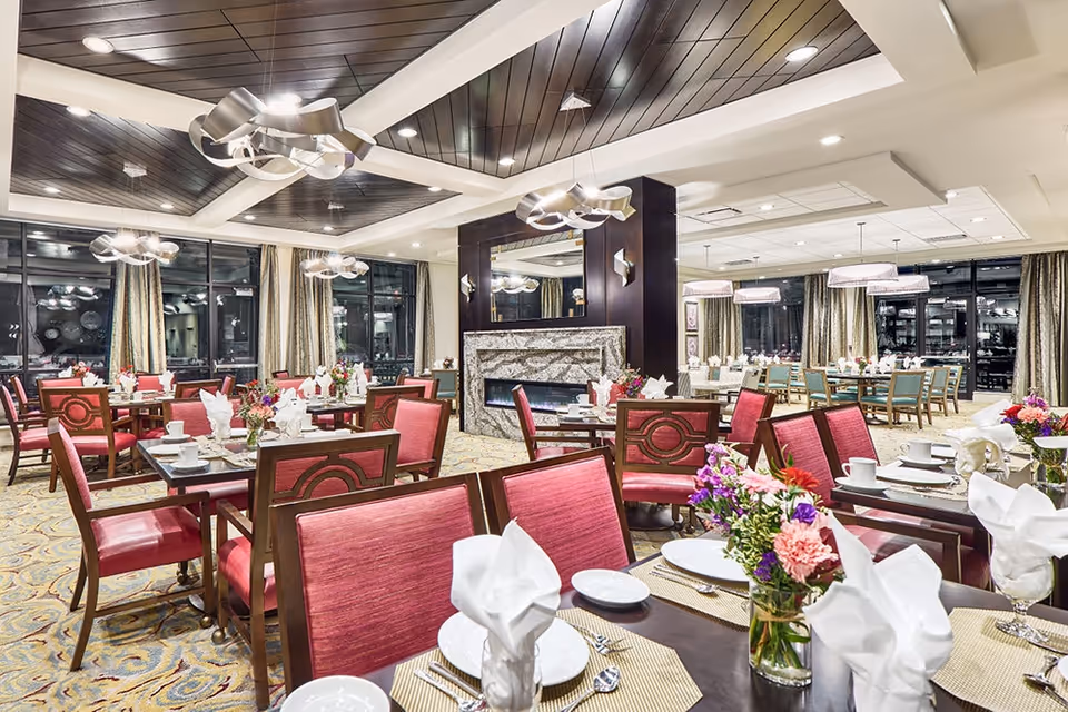 A spacious, well-lit dining room with multiple tables set for a meal. Each table has red cushioned chairs, white plates, napkins folded elegantly, silverware, and small floral centerpieces. The room features large windows with curtains, modern ceiling lights, and a decorative fireplace with a mirror above it.