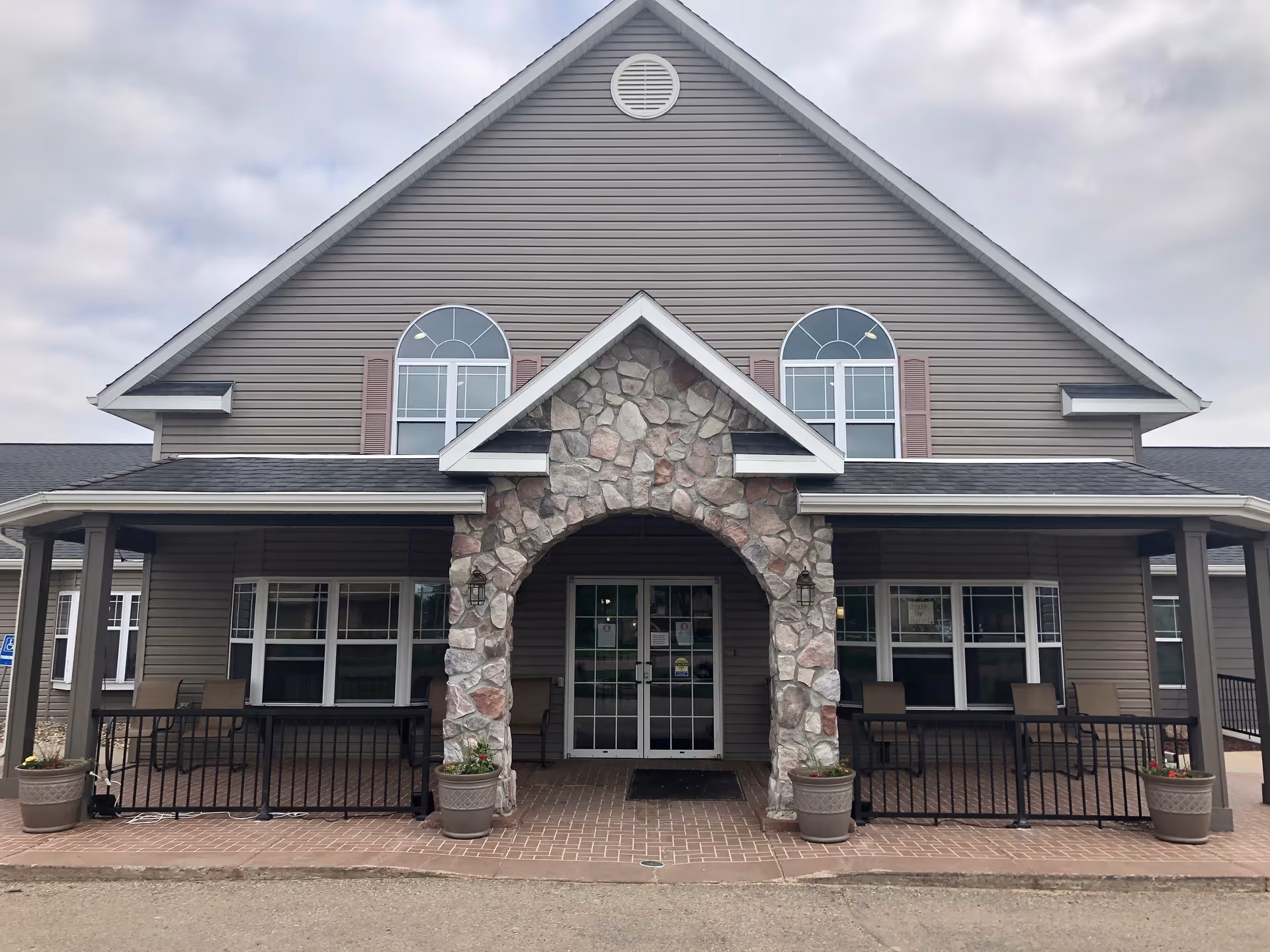 Front exterior view of StoneyBrook Suites Assisted Living building with a stone arch entrance, two large windows with brown shutters, and a covered porch area with chairs and potted plants.