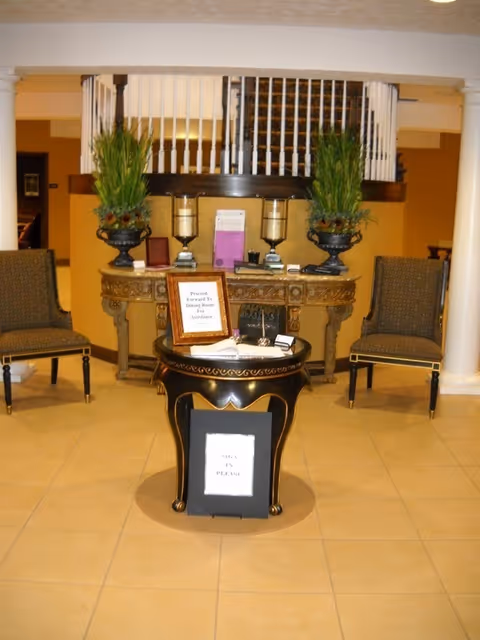 An interior lobby area with a small round table in the center holding a sign that reads 'Sign In Please.' Behind the table is an ornate console table with two large potted plants, two candle holders, and informational materials. Two upholstered chairs are positioned on either side of the console table. A staircase with white railings is visible in the background.