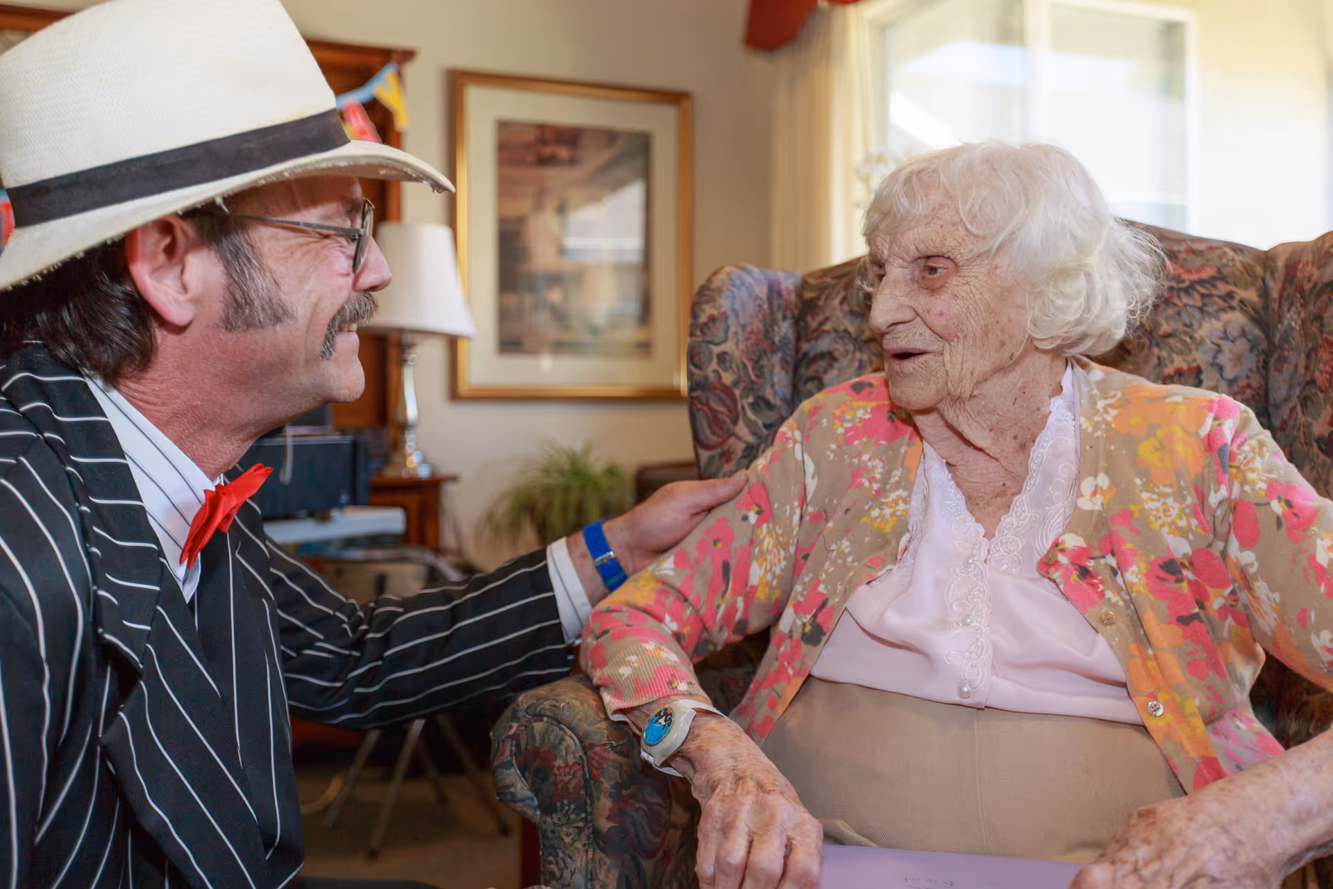An elderly woman with white hair wearing a floral cardigan and a pink blouse sits in a patterned armchair, smiling and talking with a man in a pinstripe suit, red bow tie, and white hat who is kneeling beside her with a hand on her shoulder in a warmly lit room.