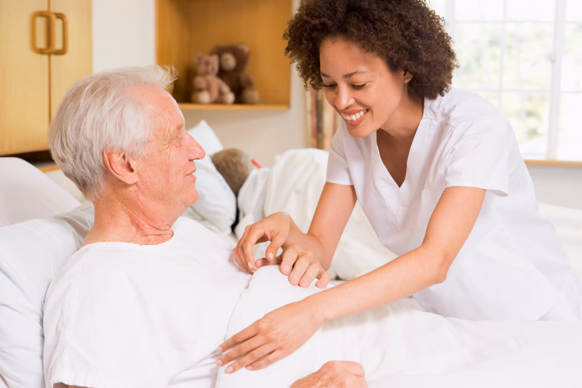 A caregiver helps an elderly man sit up in a hospital-style bedroom.