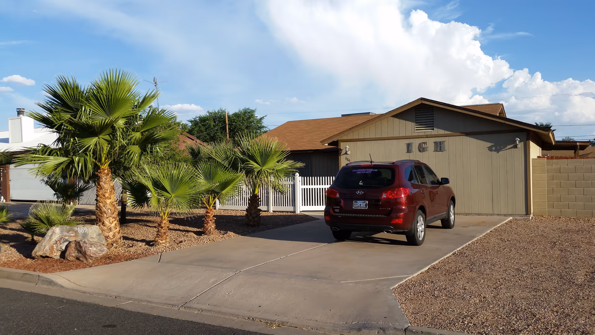 Single-story building labeled 'IGH' with a driveway, a red SUV parked in front, and palm trees with gravel landscaping under a blue sky.