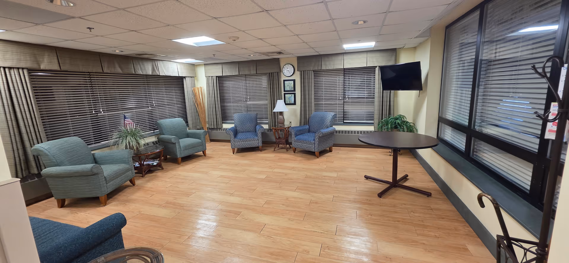 Senior living lounge with blue chairs, tables, blinds, a lamp, and a wall-mounted TV.
