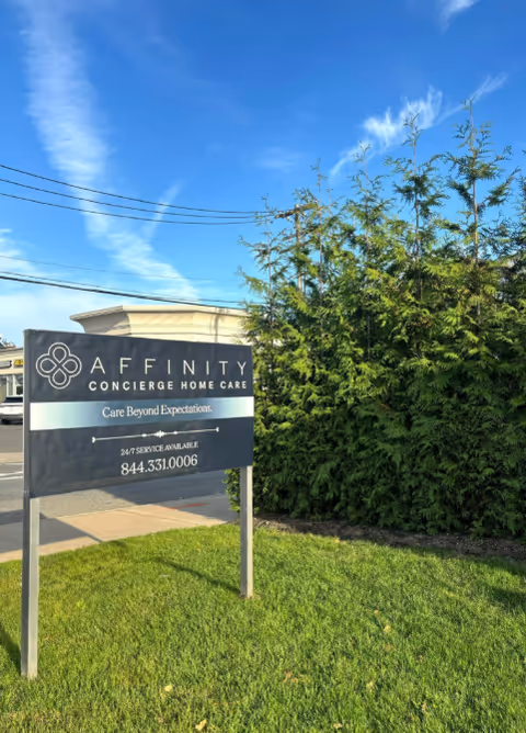 Outdoor sign reads Affinity Concierge Home Care in Hewlett, with hedge and blue sky.