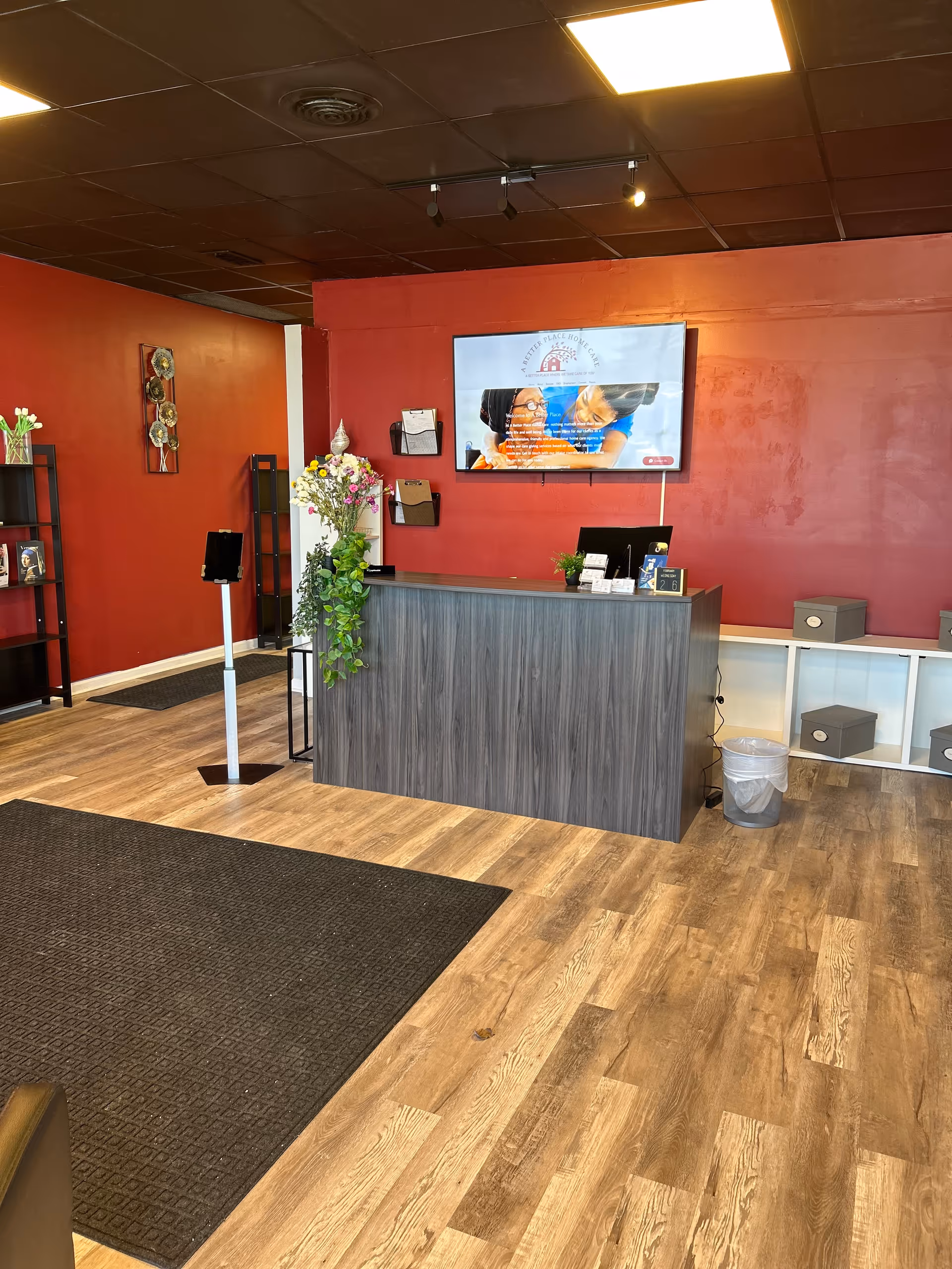Lobby reception area with a dark wood front desk, mounted TV, plants, and a red wall.