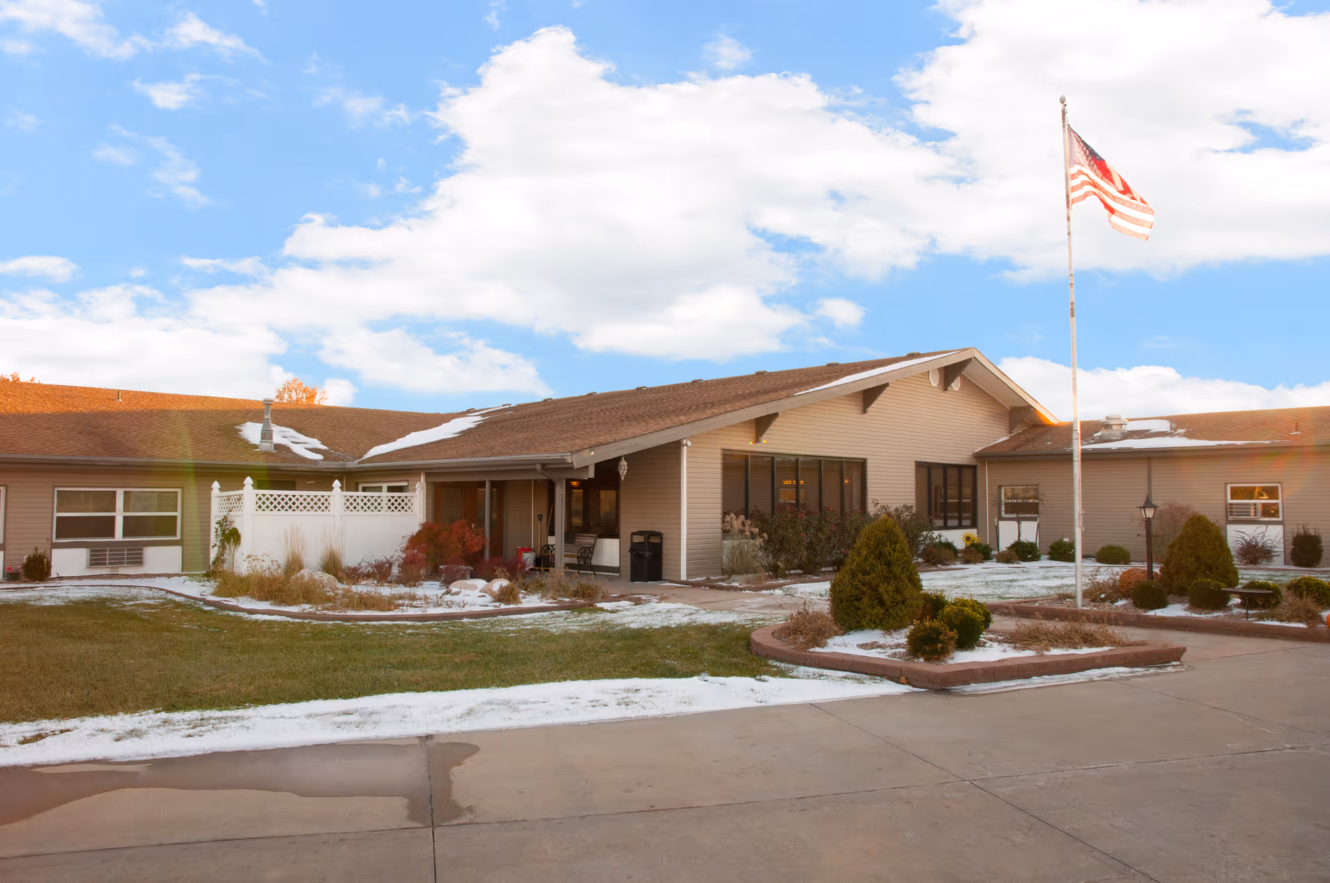 Front exterior of a single-story senior living facility with an American flag, landscaped beds, and light snow on the ground.