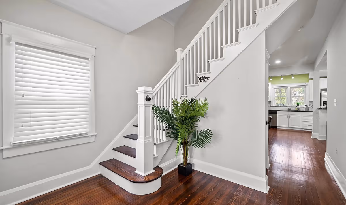 Bright interior foyer with staircase, white railings, hardwood floors, and kitchen visible.
