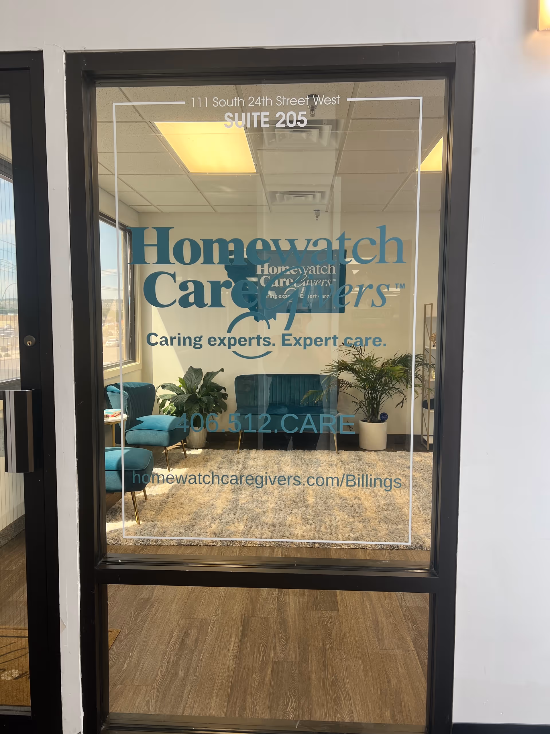 Glass door reveals a waiting lobby with teal chairs and potted plants inside.