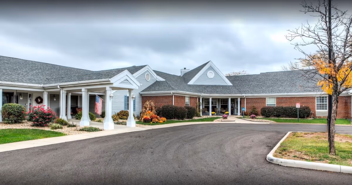 Single-story brick and siding senior living facility front entrance with a covered porte-cochère, landscaping, and an American flag.