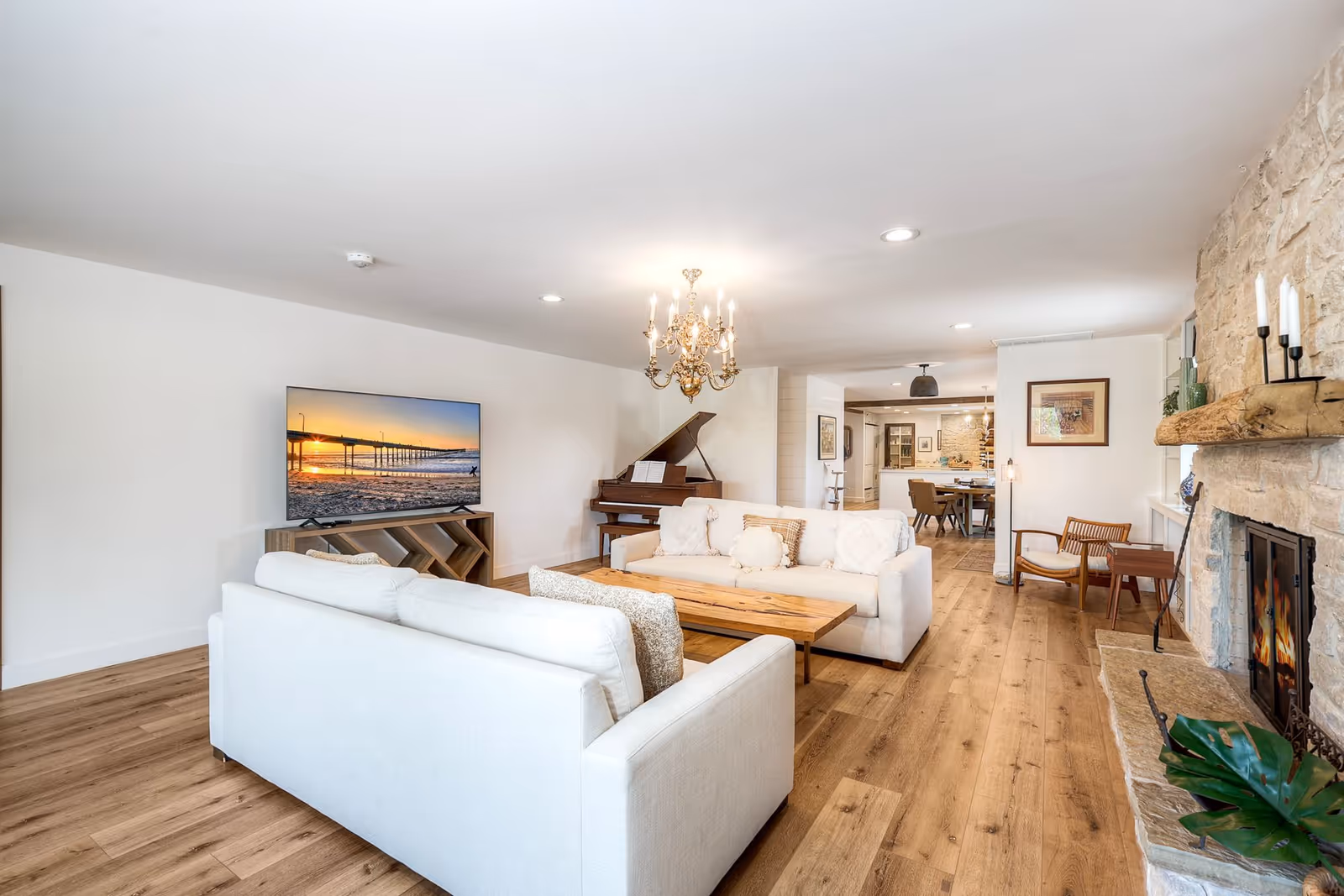 Bright living room with white sofas, chandelier, fireplace, piano, and wooden floors.
