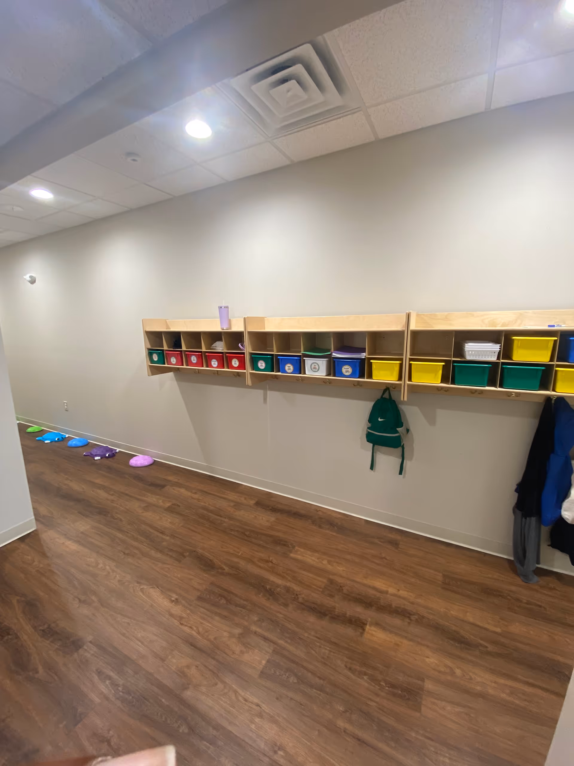Empty activity room with wall-mounted cubbies, colorful bins, and coats on hooks.