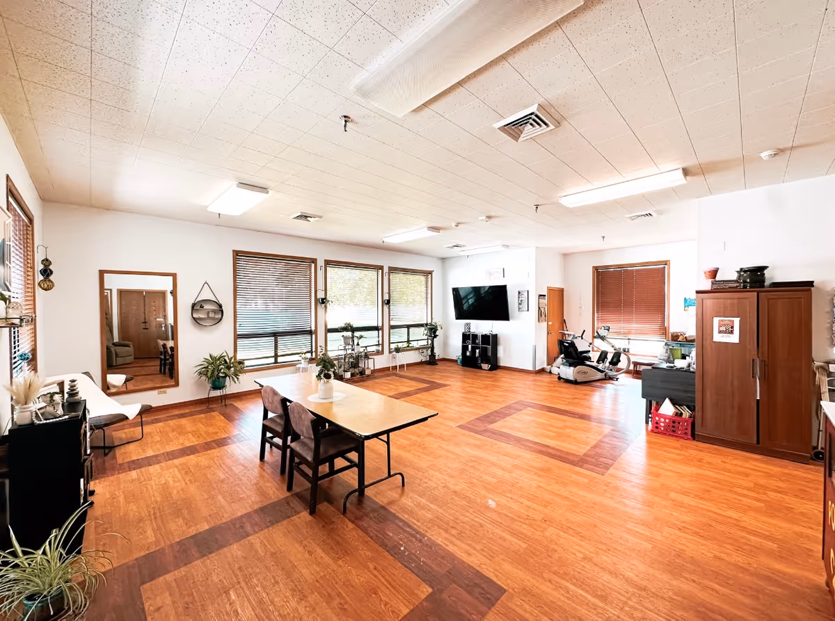 Spacious senior living common room with table, chairs, plants, windows, and exercise area.