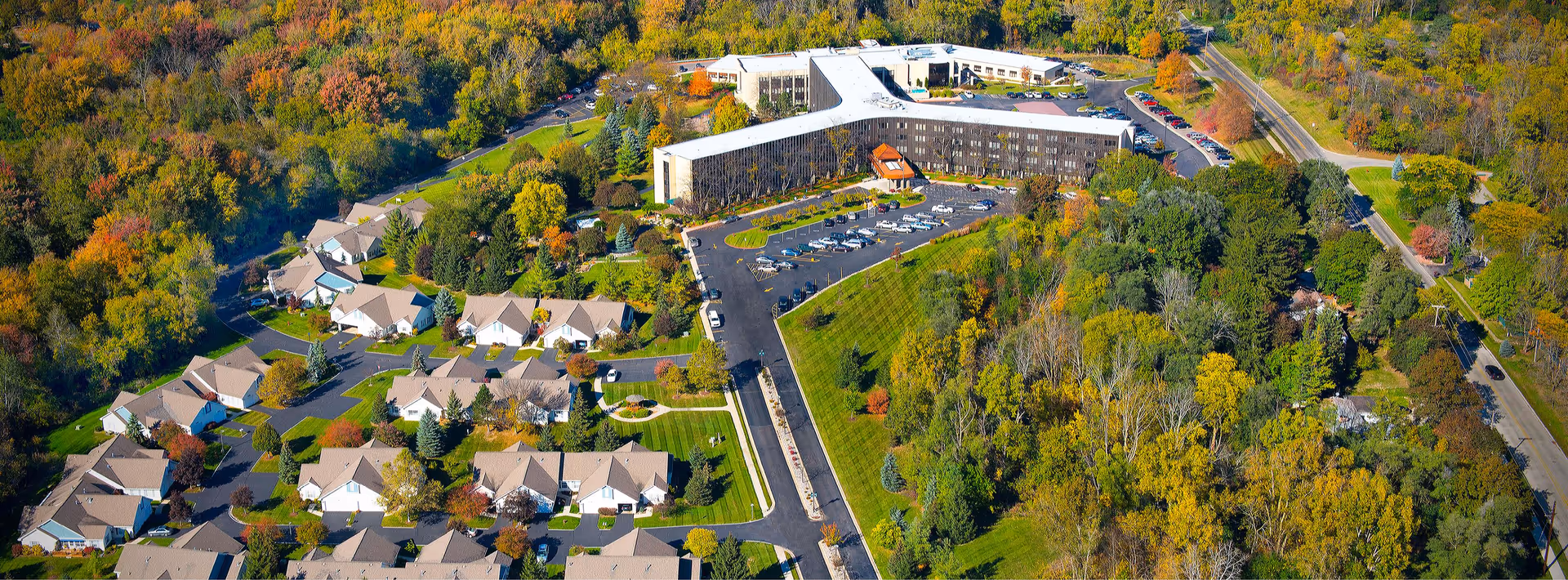 Aerial view of Burcham Hills senior living facility surrounded by trees with autumn foliage. The image shows a large main building with a parking lot and several smaller residential buildings arranged in a neighborhood-like layout. Roads and green spaces are visible throughout the property.