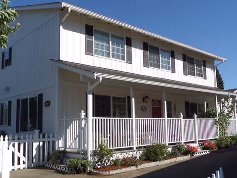 Exterior view of a two-story white senior home with a covered porch, white railing, and a red front door. The house has multiple windows with shutters and is surrounded by small bushes and flowers along the front walkway.