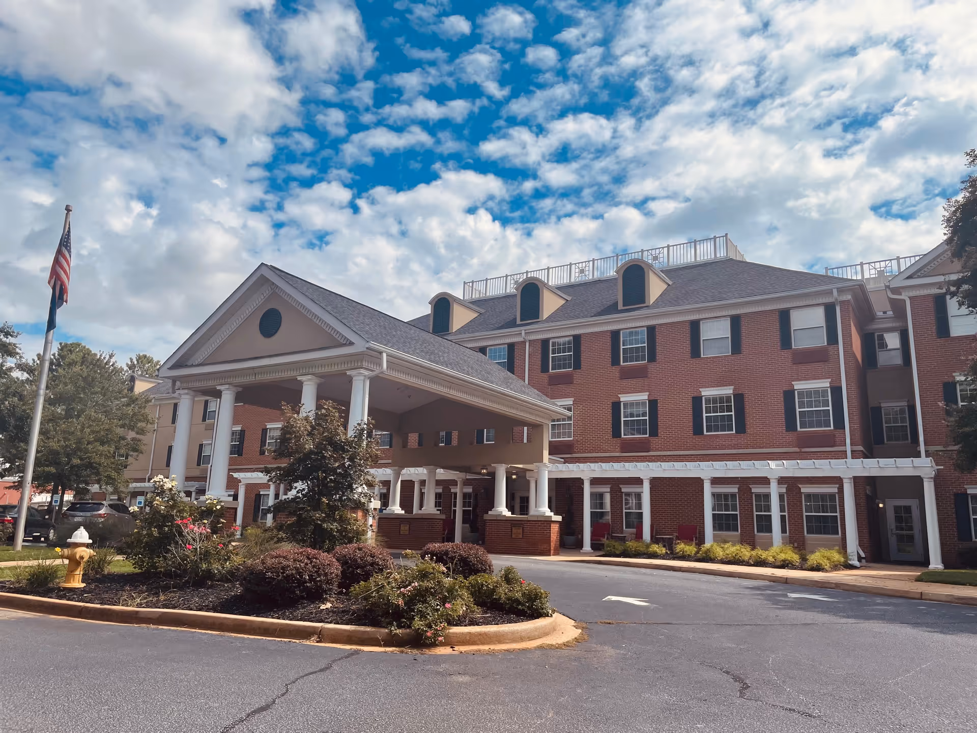 Exterior view of a multi-story brick building with white columns and a covered entrance. The building is surrounded by landscaped bushes and trees, with an American flag on a flagpole to the left. The sky is partly cloudy with patches of blue.