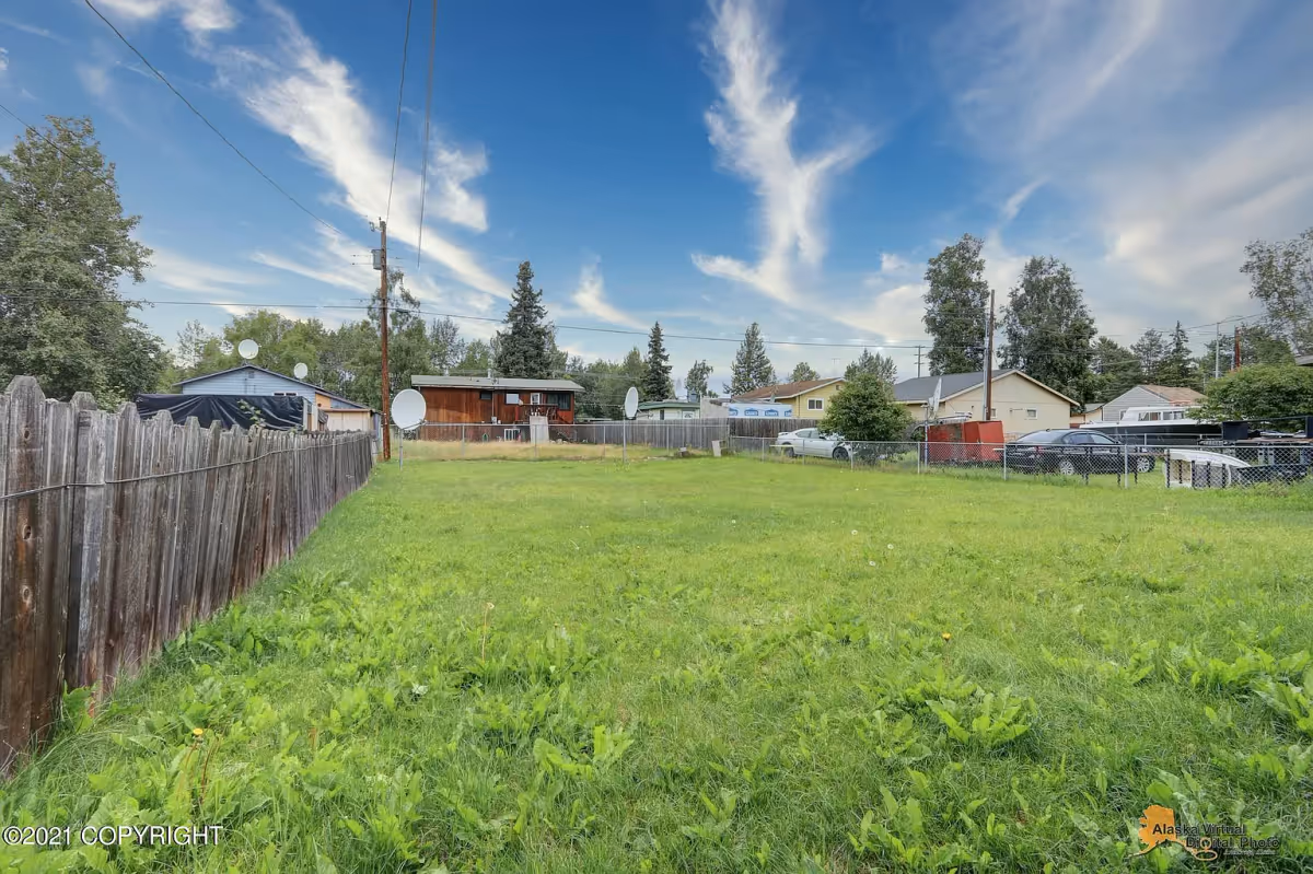 A grassy fenced yard with a wooden fence on the left and chain-link fences on the right and in the background. Several houses and trees are visible beyond the fences under a blue sky with wispy clouds.