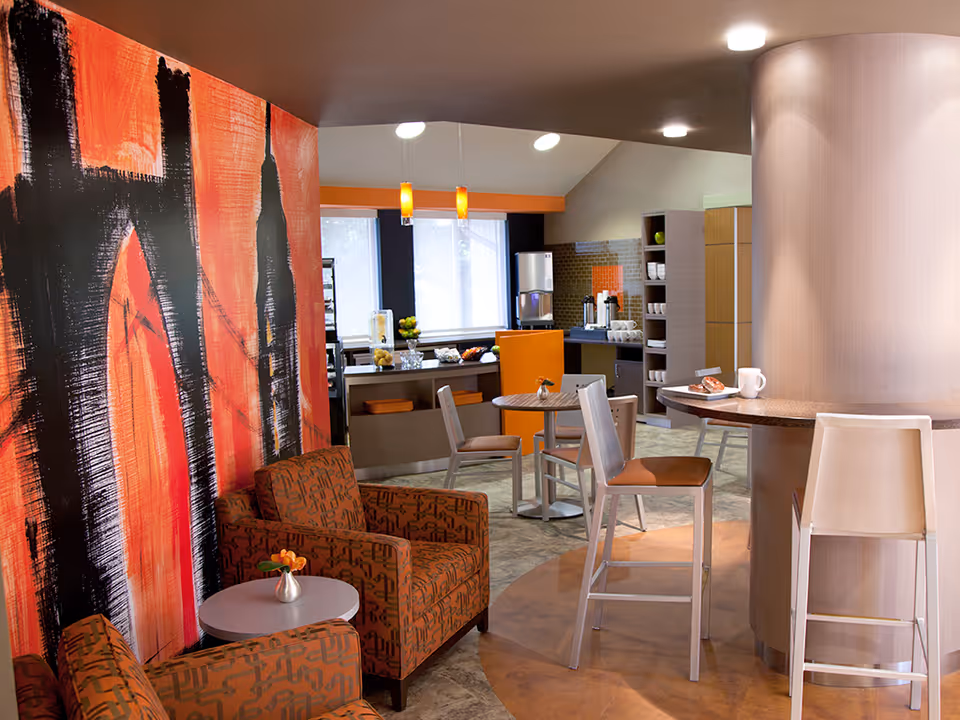 A modern common area in a senior living facility featuring two patterned armchairs with a small round table between them, a curved counter with two high chairs, and a kitchenette in the background with a coffee machine and shelves. The wall on the left has a large abstract mural in orange and black tones, and the room is well-lit with ceiling lights and pendant lamps.