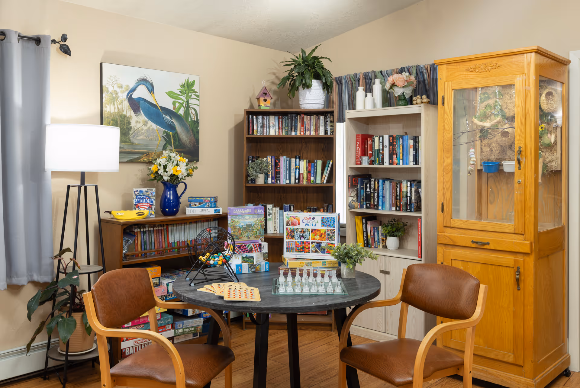 Cozy senior living activity room featuring a round table, chairs, bookshelves, plants, and a wooden cabinet.