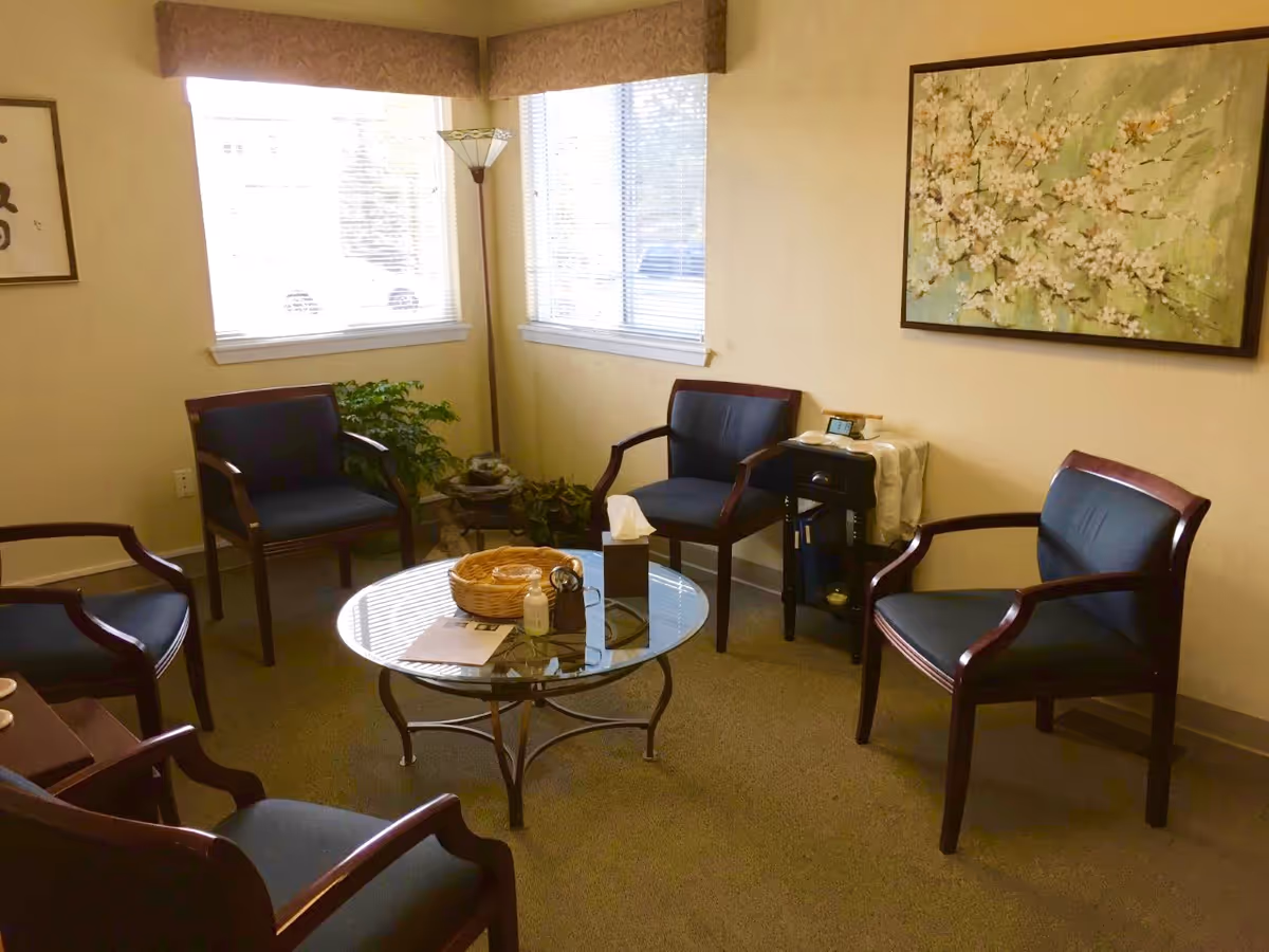 Senior living lounge with glass coffee table, multiple chairs, plants, and wall art.