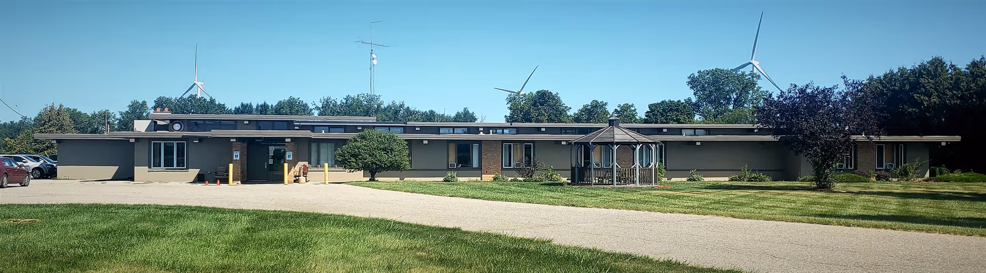Wide exterior view of a single-story senior living facility building with a flat roof, surrounded by green grass and trees. There is a small gazebo on the lawn in front of the building, a paved driveway leading to the entrance, and wind turbines visible in the background.