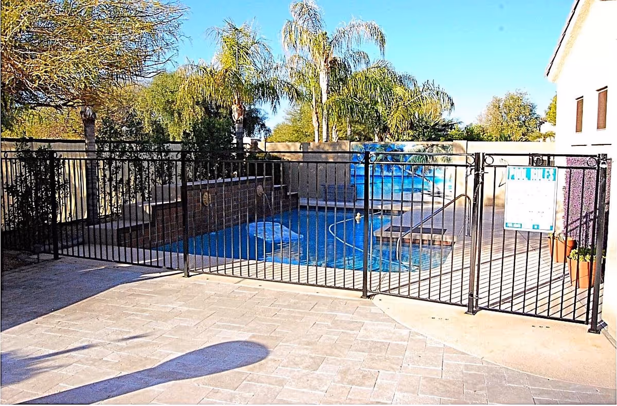 Outdoor swimming pool area enclosed by a black metal fence with a gate. The pool has water features on the far side and a mural of palm trees on the back wall. There are palm trees and other greenery surrounding the pool area under a clear blue sky.