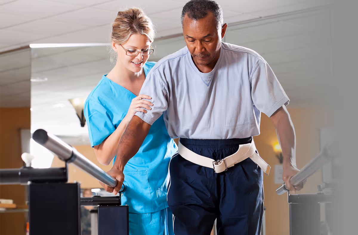 A healthcare professional in blue scrubs assists an elderly man using parallel bars for walking rehabilitation in an indoor therapy setting.