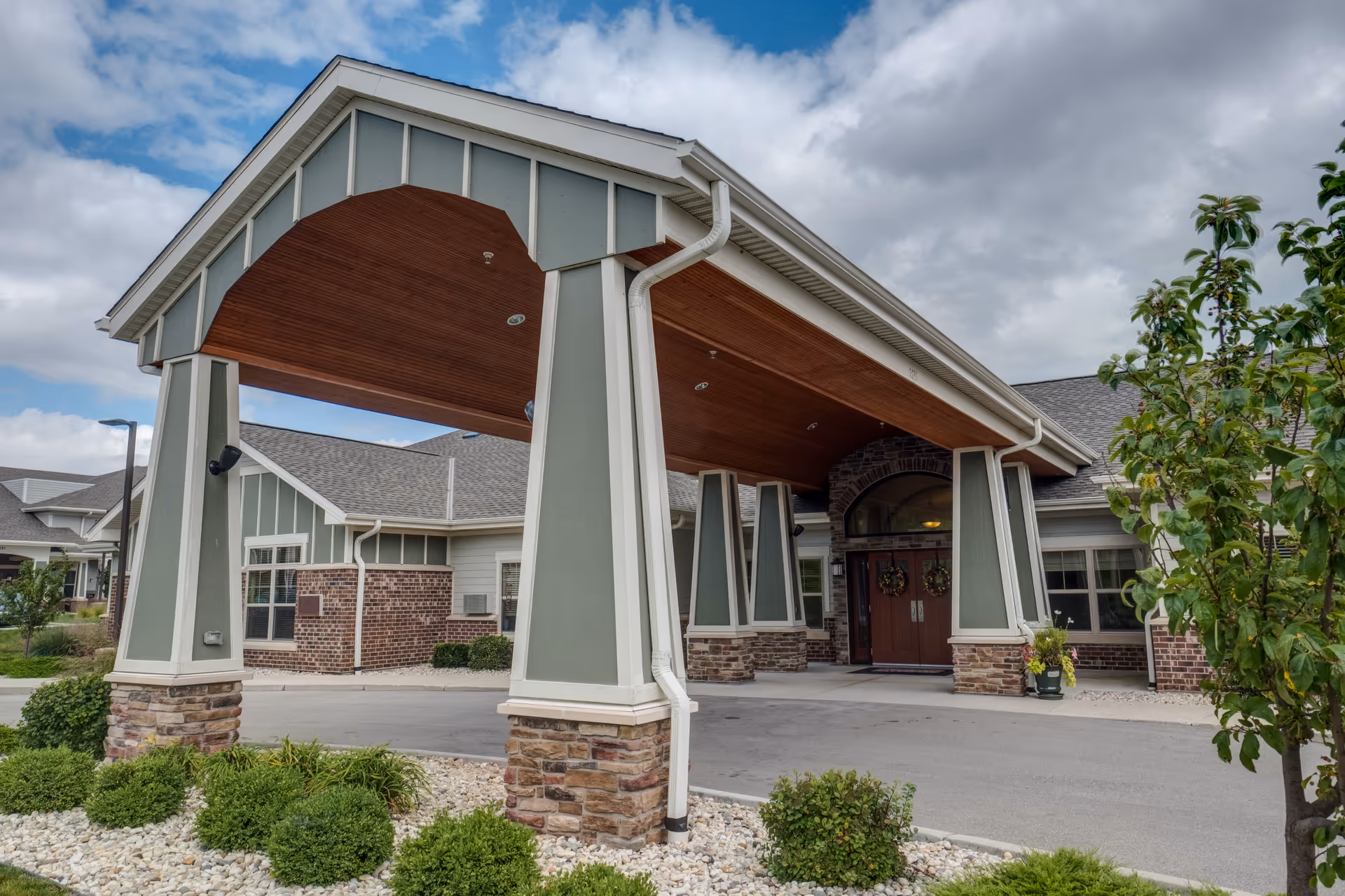 Entrance of Heritage Court Memory Care facility featuring a covered driveway supported by large pillars with stone bases, a wooden ceiling, and double doors decorated with wreaths. The building has a combination of brick and light green siding with white trim, surrounded by landscaping with bushes and a tree under a partly cloudy sky.