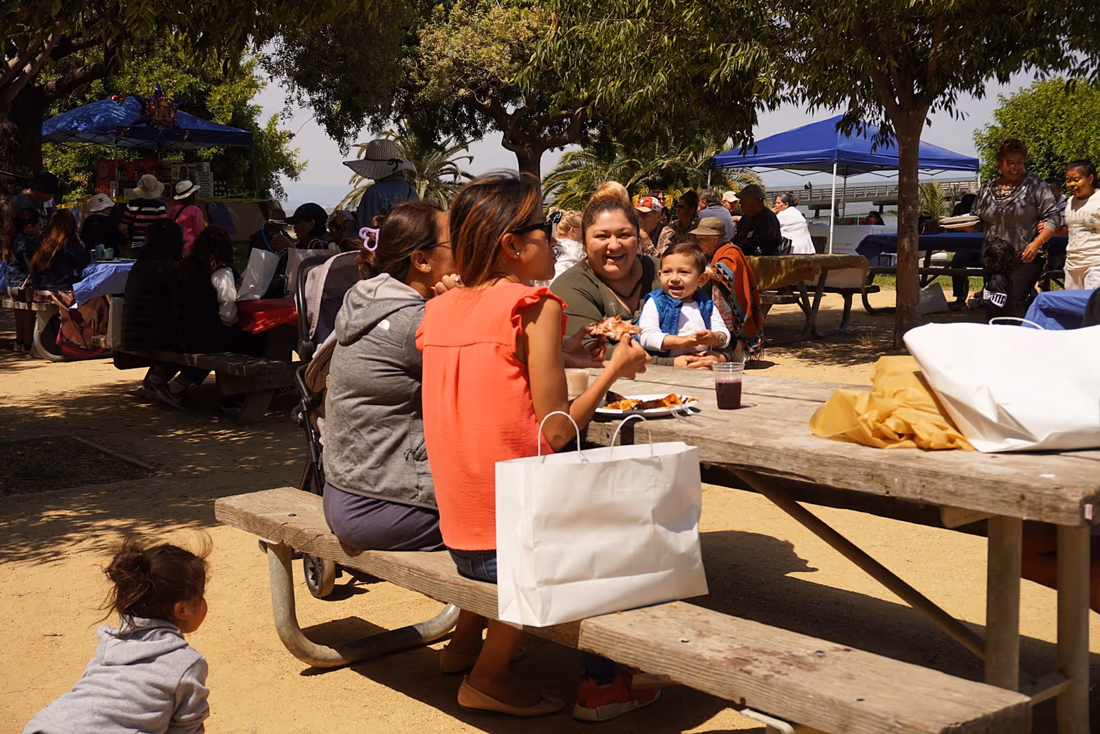 People gather at outdoor picnic tables under trees for a communal outdoor gathering.