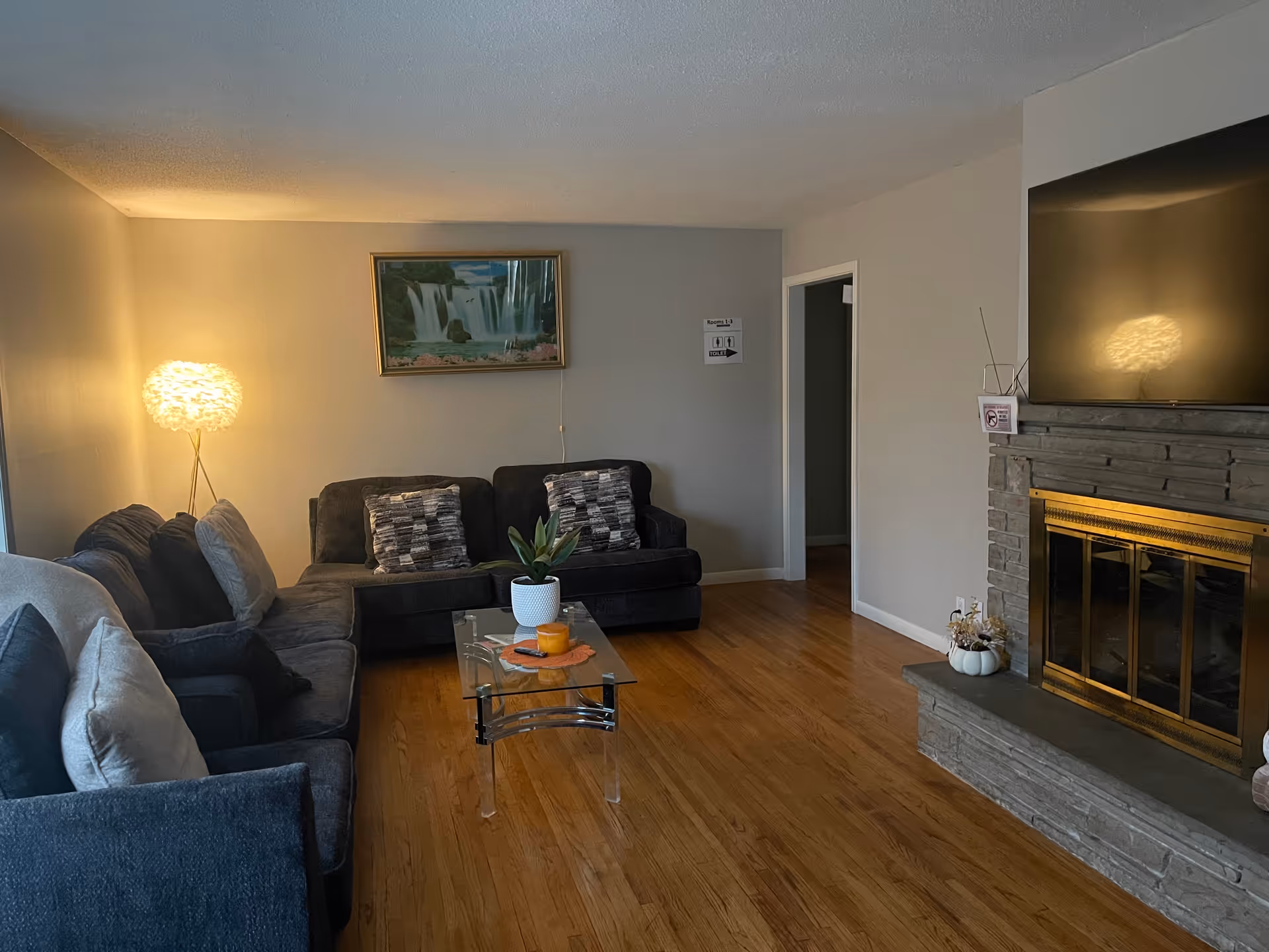 A cozy living room with a sectional sofa, glass coffee table, lamp, painting, and fireplace.