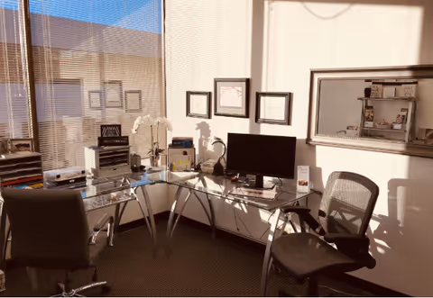 An office space with glass desks, chairs, computer, and framed certificates.