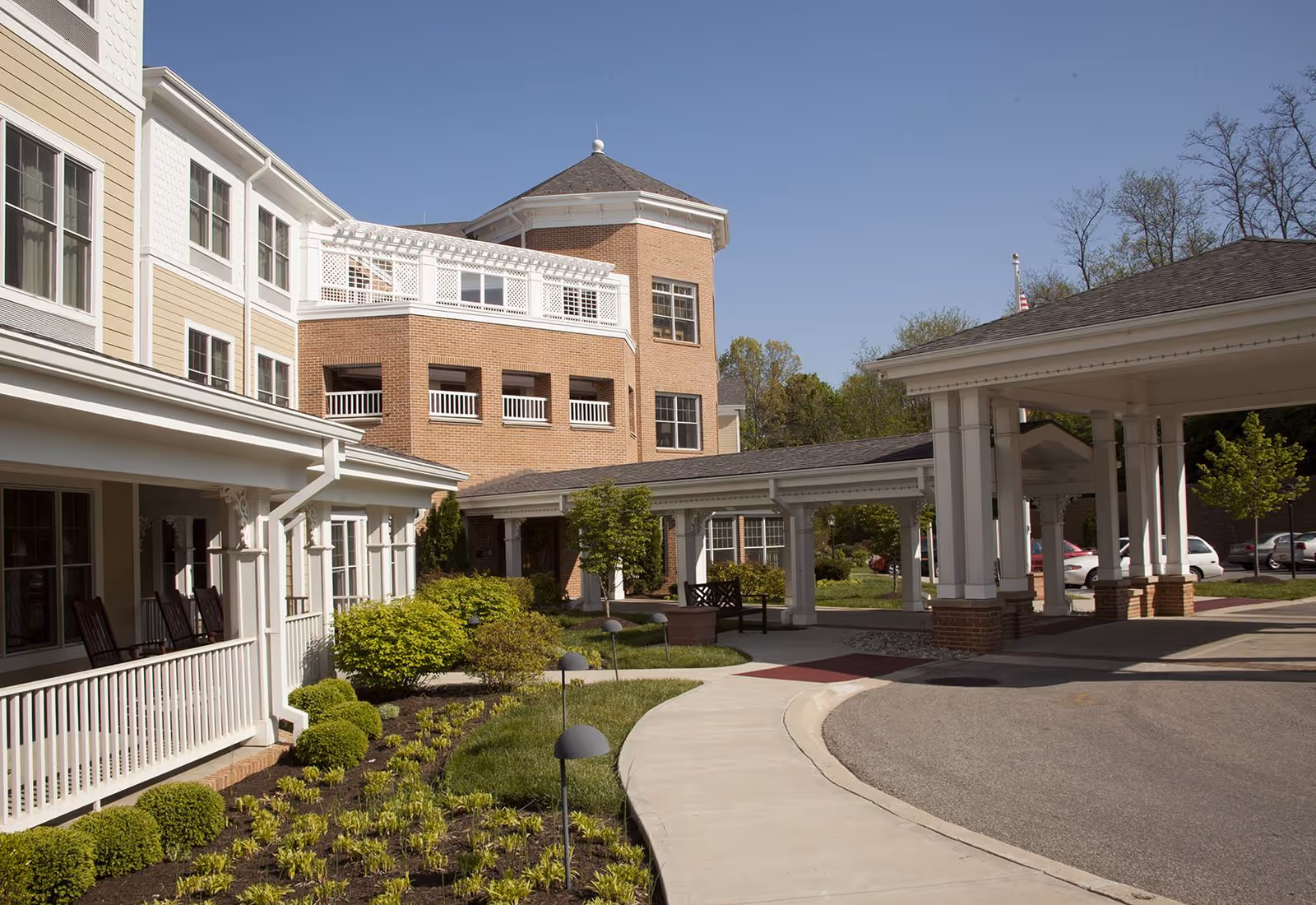 Front exterior of a senior living facility showing a covered entrance/porte-cochere, curved driveway, and landscaped walkway.