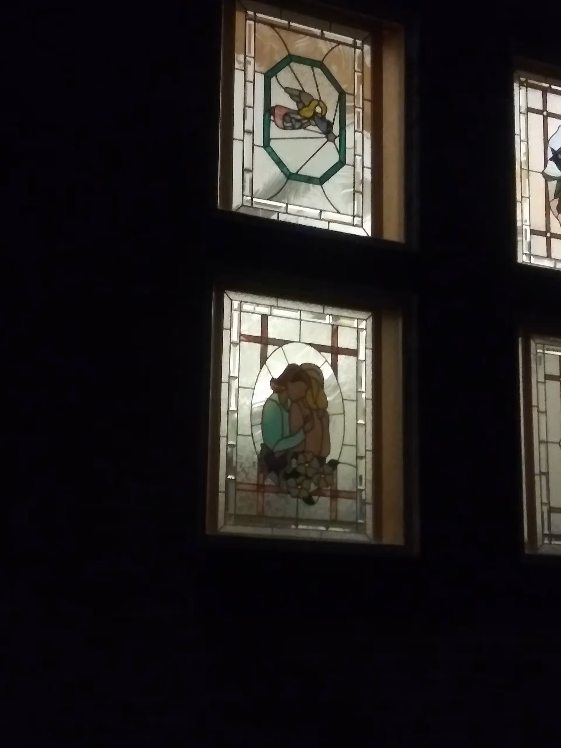 Dark exterior wall with illuminated stained-glass window panels featuring figures.