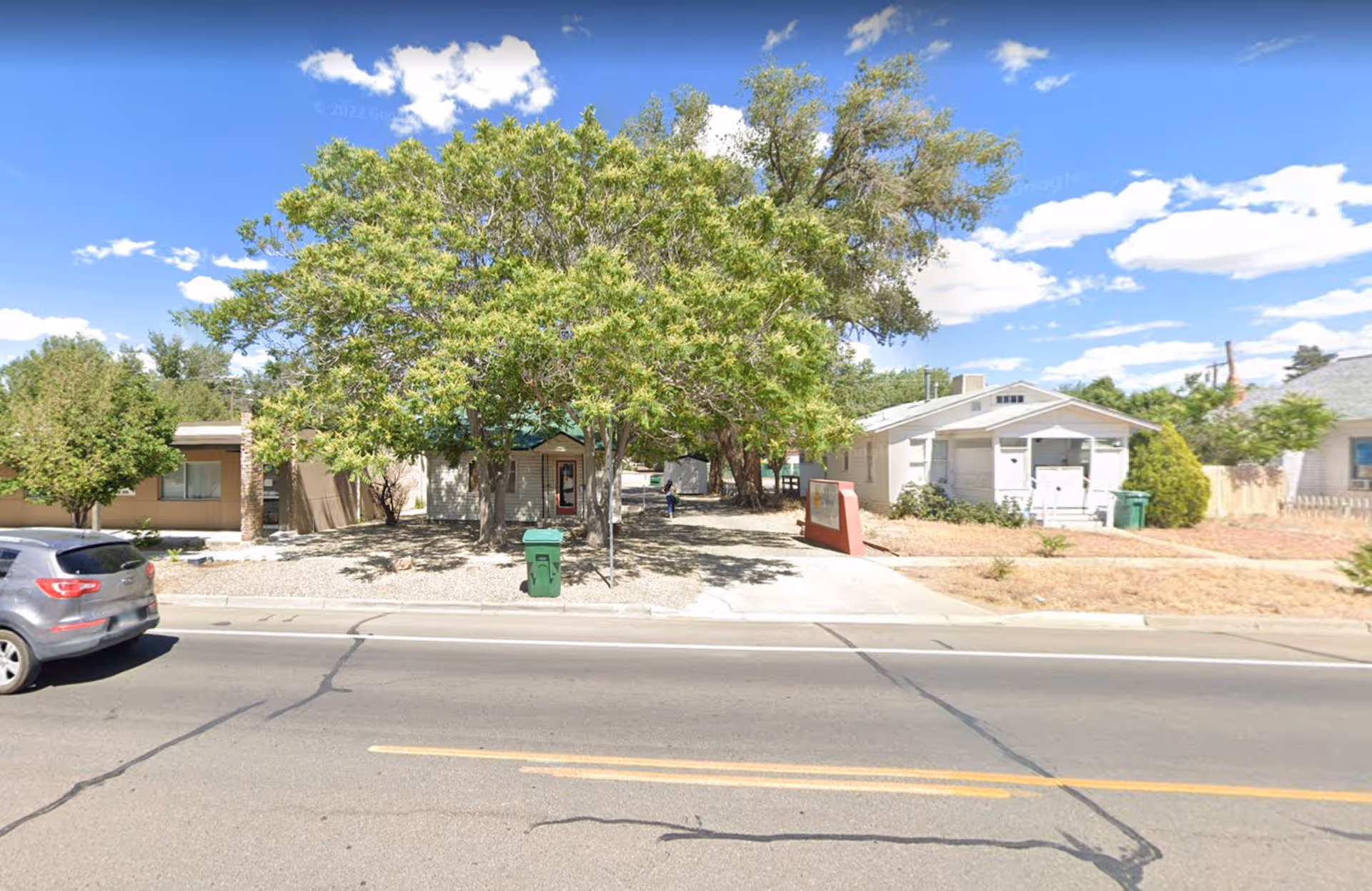 Front view of a small building with large trees and a sidewalk along the street.