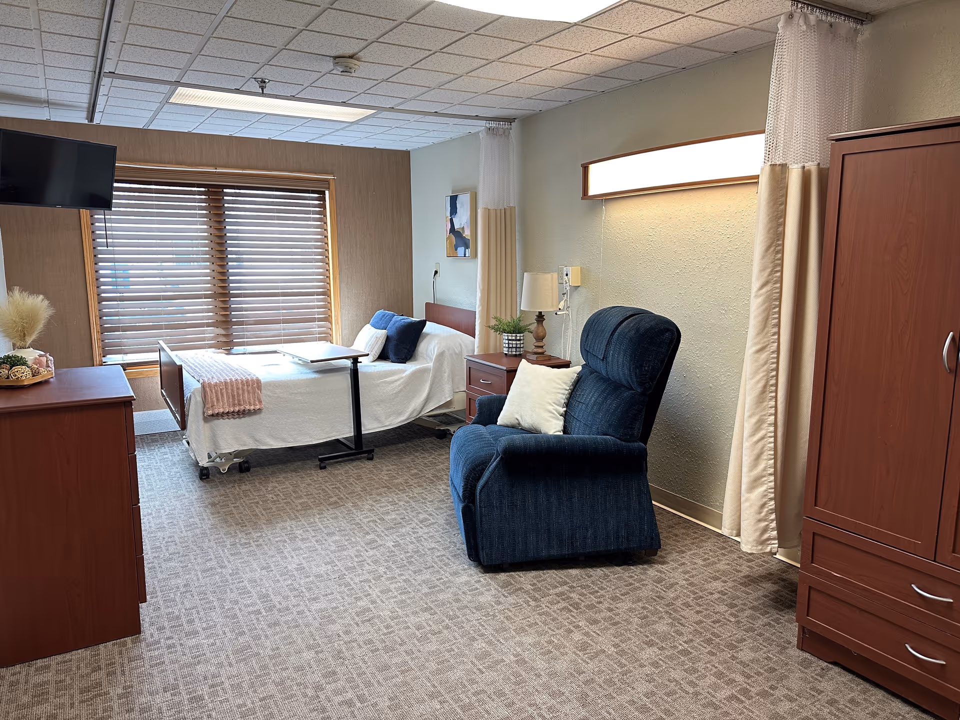 A well-lit rehabilitation center room featuring a hospital bed with white linens and a pink throw blanket, a blue recliner chair with a white pillow, a wooden dresser, a wooden nightstand with a lamp and a small plant, a wall-mounted TV, and a large window with wooden blinds. The room has beige carpet and light-colored walls with a privacy curtain partially drawn.