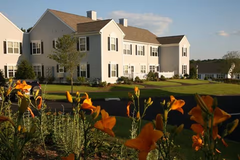 A large, two-story white building with multiple windows and a sloped roof, surrounded by a well-maintained lawn and orange flowers in the foreground under a partly cloudy sky.