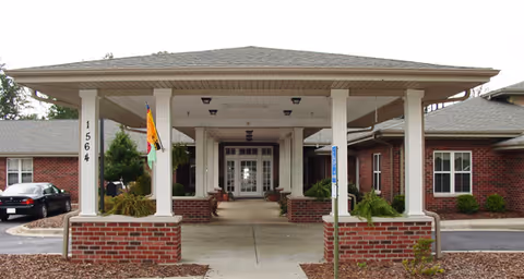 Entrance of a single-story brick building with a covered driveway supported by white columns. The building has a gray shingled roof and several windows. There is a flag hanging from one of the columns and a handicap parking sign near the entrance.