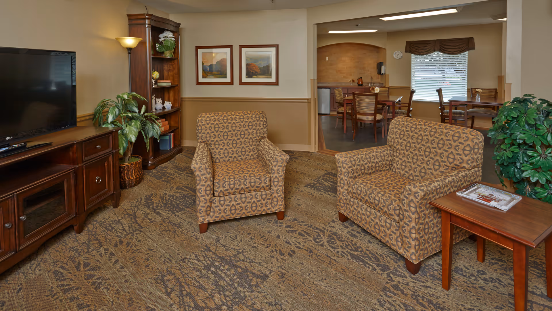 Cozy common room with two patterned armchairs facing a TV on a wooden cabinet, bookshelf, plants, and a dining area visible through an open doorway.