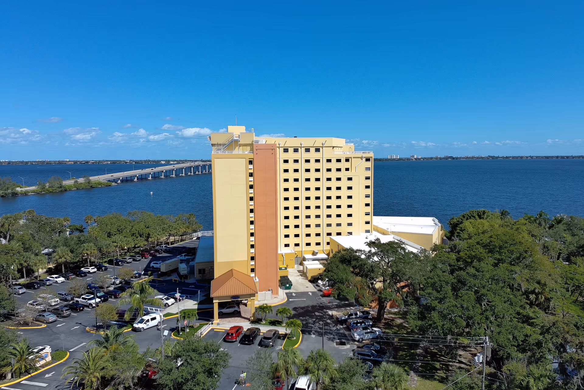 A tall tan senior living building by a large blue body of water with parked cars.