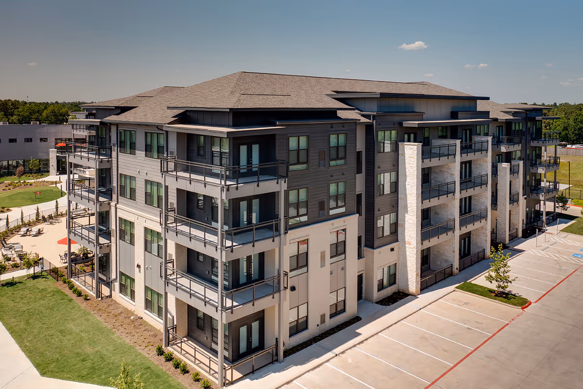 Exterior view of a modern multi-story senior living facility building with balconies, surrounded by a parking lot and landscaped green areas under a clear sky.