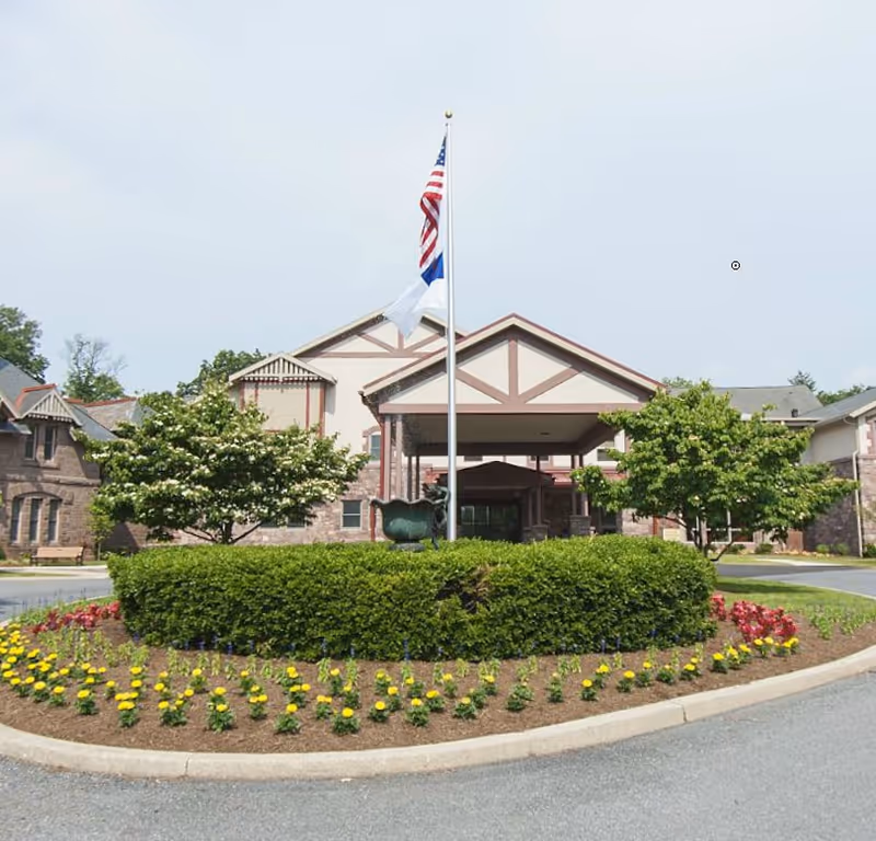 Front exterior view of Cornwall Manor building with a circular driveway, a flagpole displaying the American flag and another flag, surrounded by neatly trimmed bushes and colorful flower beds.