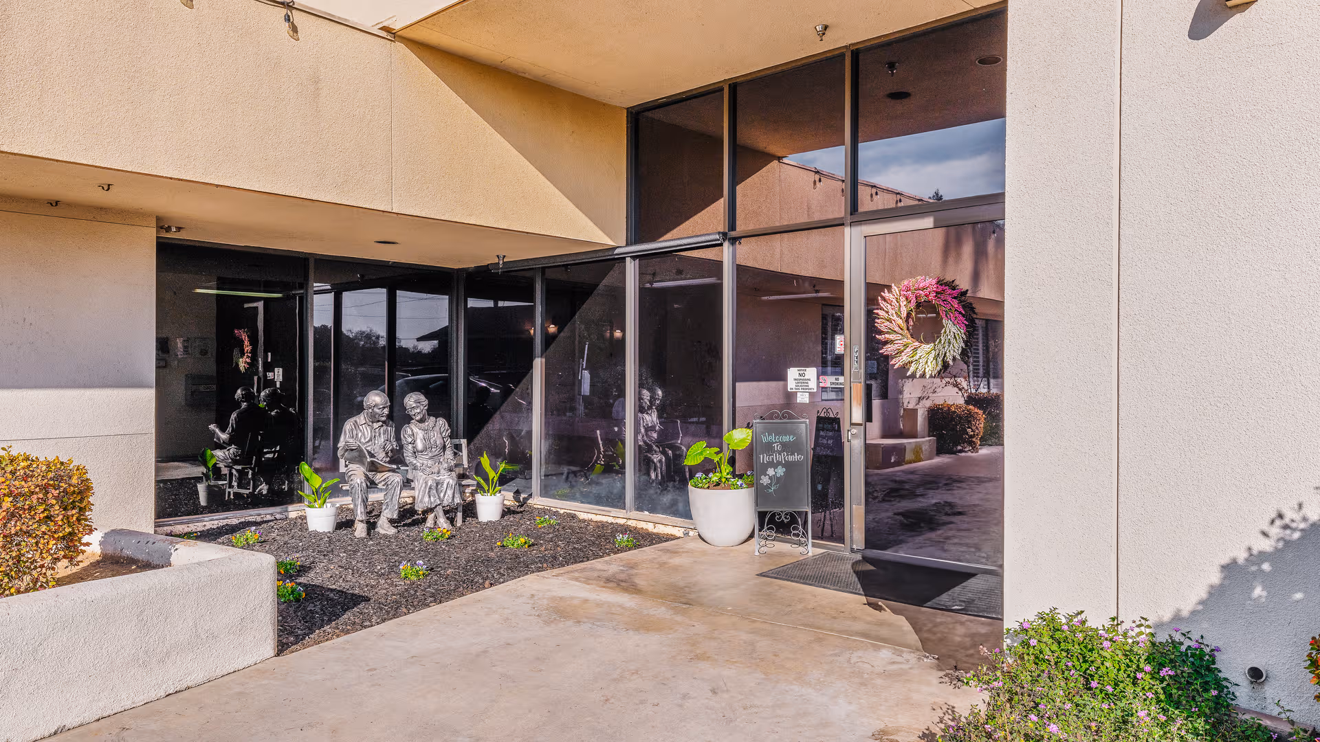 Entrance to North Point Healthcare & Wellness Centre with glass doors, planters, wreath, and seated statues.